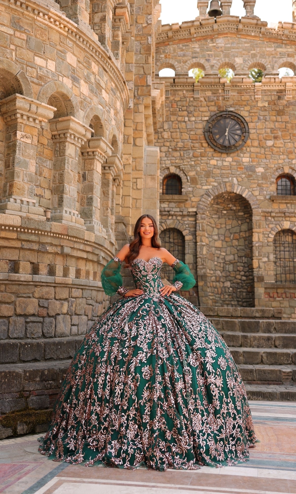 A woman in the Amarra 54238 Sequin Quinceanera Dress—featuring green fabric, silver embroidery, and elegant puff sleeves—stands smiling with hands on hips before a castle-like stone building.